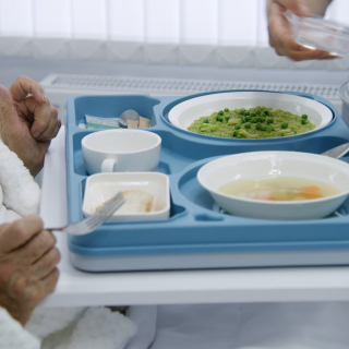 Nurse opens big lunch box with meal to female old patient. Elderly woman resting in bed in bright hospital room, recovering after successful surgery, eating food. Modern medical facility or clinic.