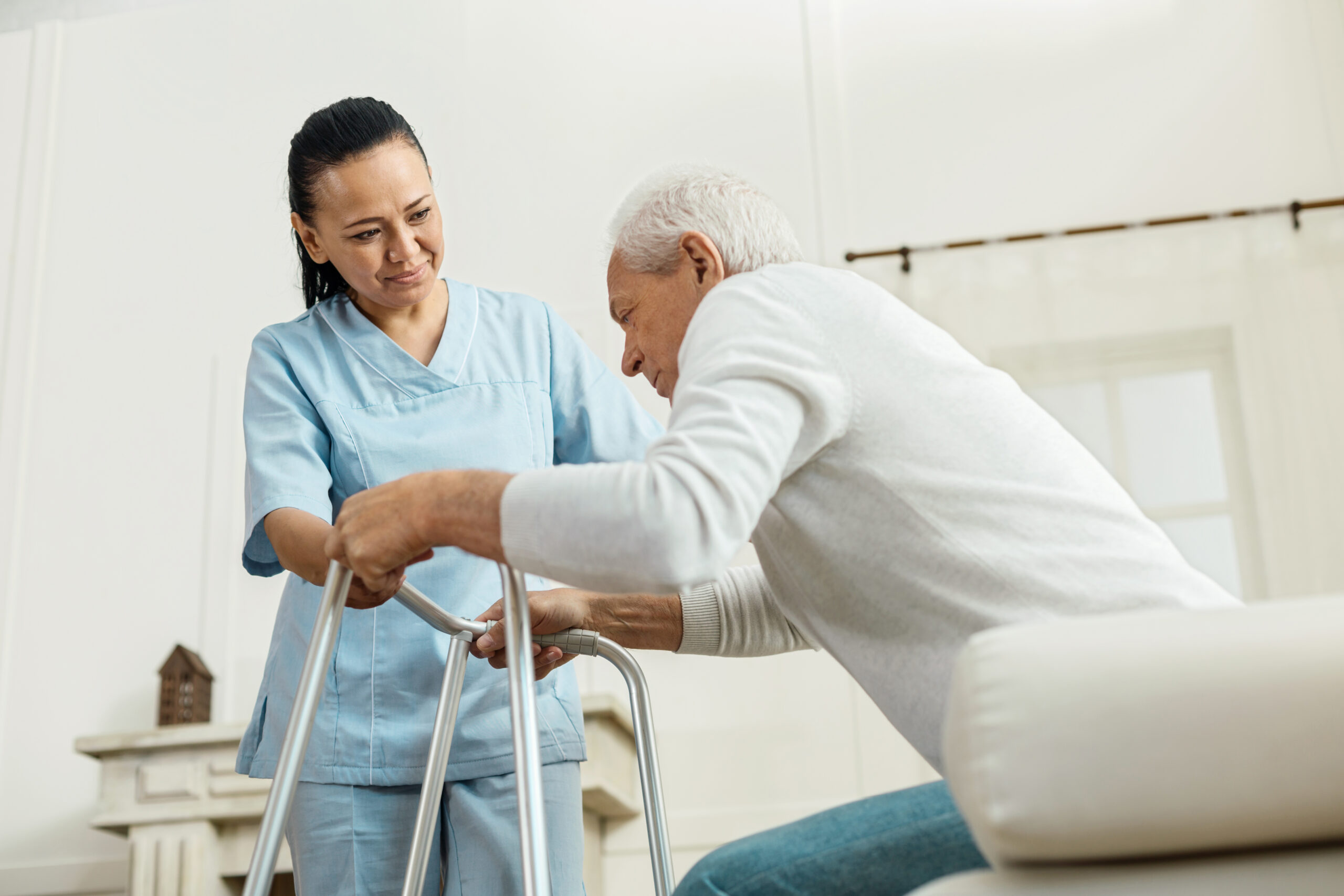 Professional caregiver. Joyful nice pleasant woman smiling and looking at the elderly man while helping him to walk